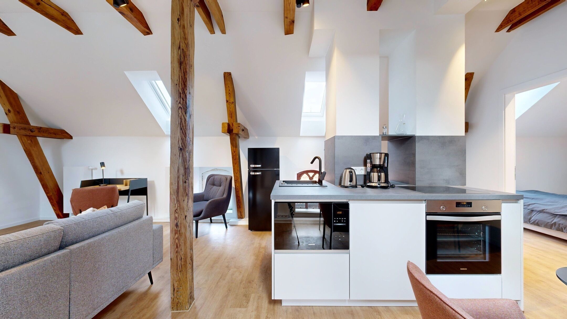 A modern loft-style apartment with exposed wooden beams, a kitchen with a dishwasher and oven, a gray sofa, and a dining area.
