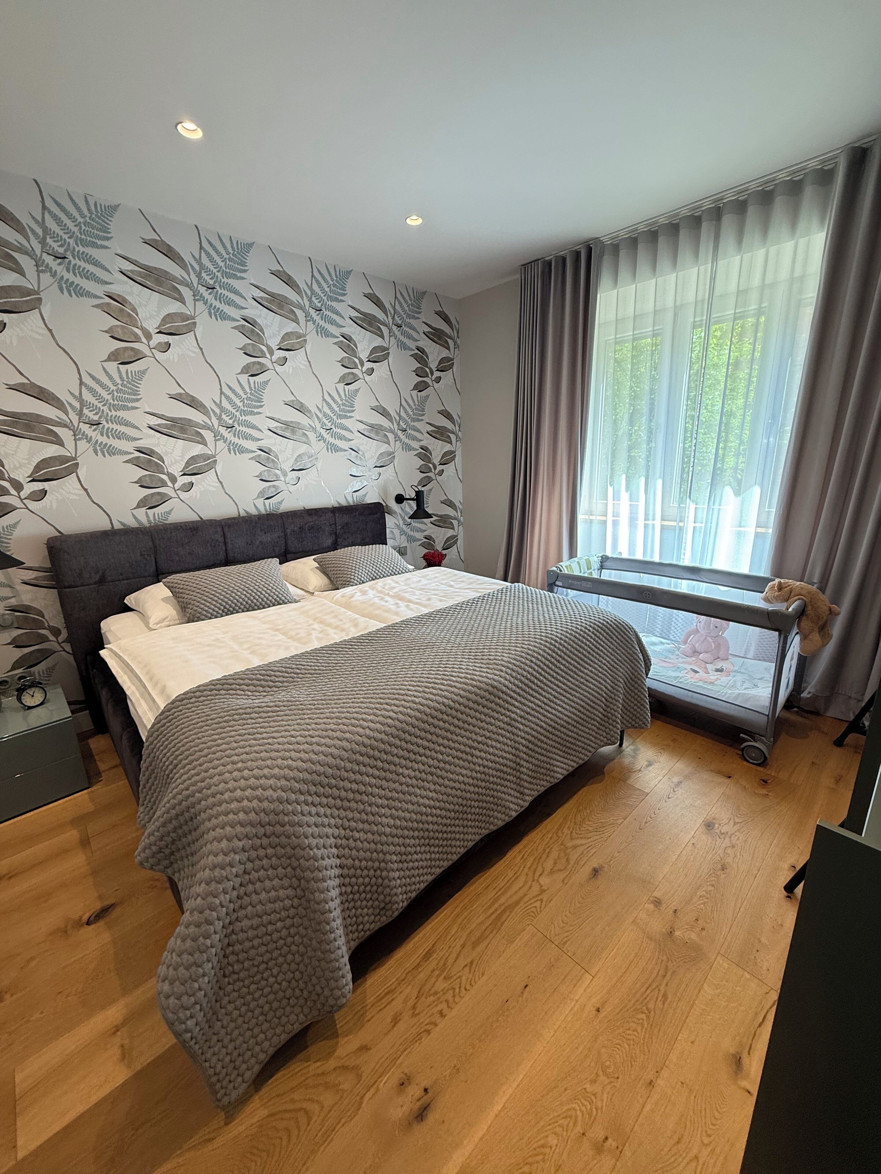 Une chambre à coucher avec un lit, une table de chevet, une fenêtre avec des rideaux et un mur orné d’un motif de feuilles.