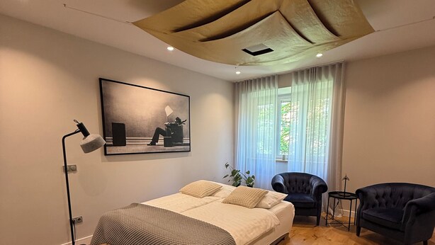 Une chambre à coucher moderne, avec un lit, deux fauteuils, une petite table, une lampe sur pied et un tableau au mur.