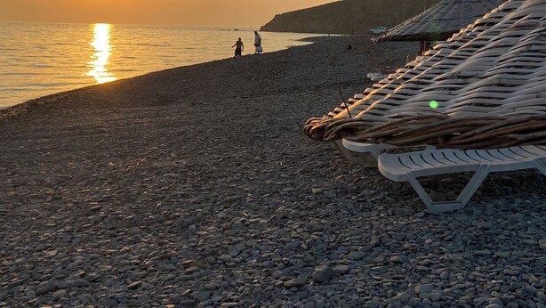 A pebbled beach with a sunset, a few people, and boats.