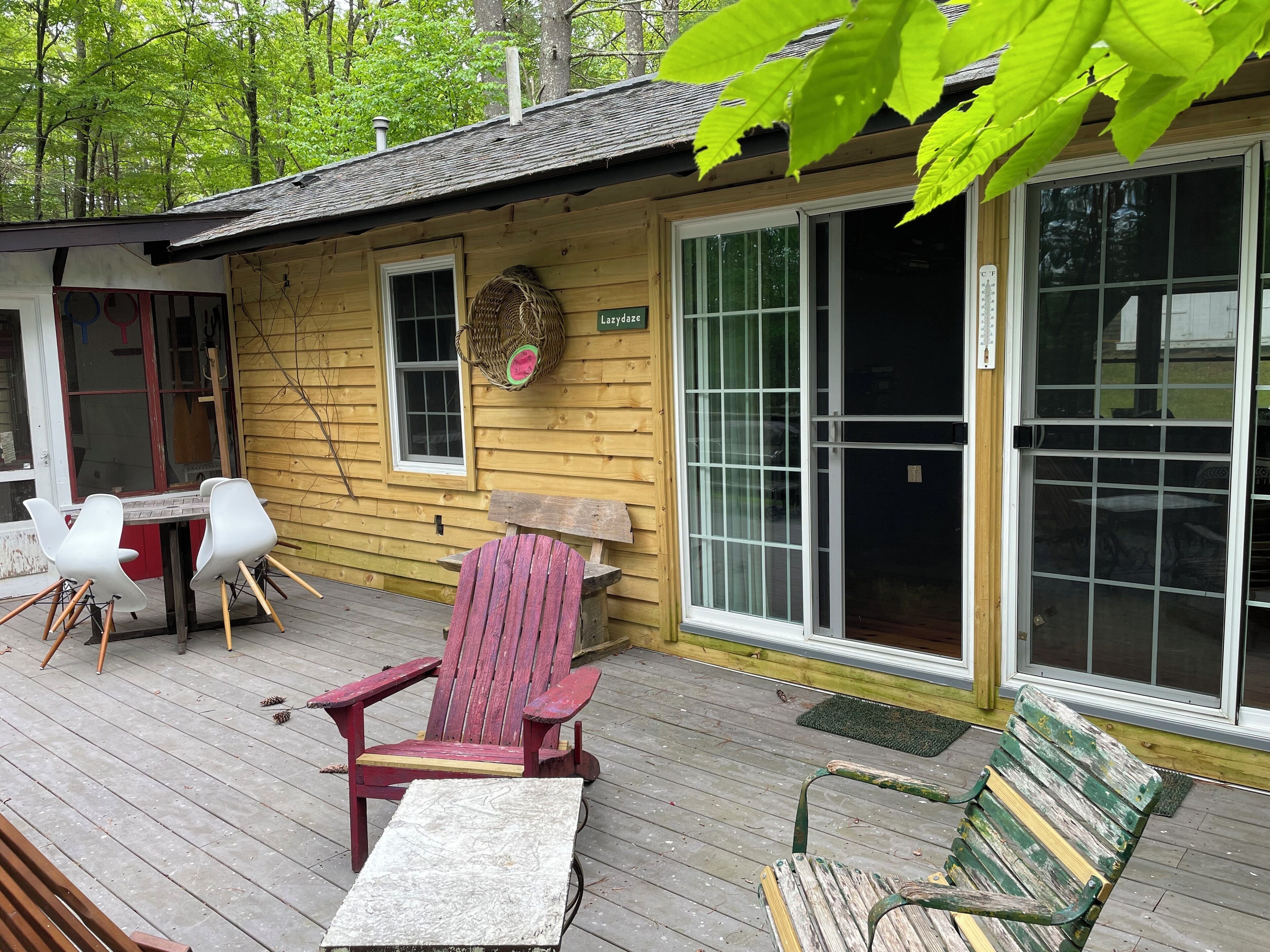 A wooden cabin with a deck, a red Adirondack chair, a white table, and a green bench.