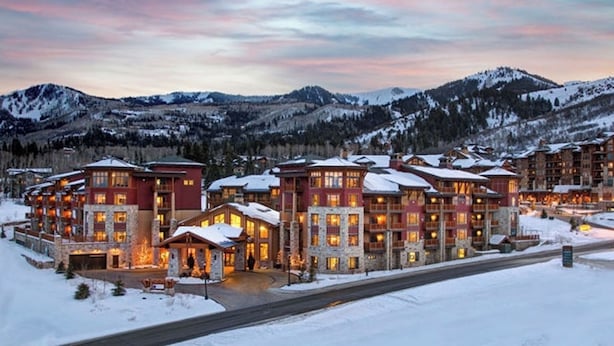 A multi-story lodge with stone and wood architecture, featuring balconies and large windows, set against a snowy mountainous backdrop.