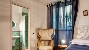 Un dormitorio con cama, mesita de noche con lámpara, una silla, ventana con cortinas y baño con lavamanos y espejo.