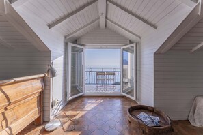 Ett smalt rum med högt i tak och utsikt över havet, en träbänk, en lampa och ett tidningsställ.