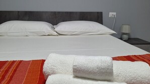 Une chambre d’hôtel avec un lit, des oreillers blancs, une couverture rayée rouge et orange, et un tableau représentant des mains formant un cœur au-dessus du lit.