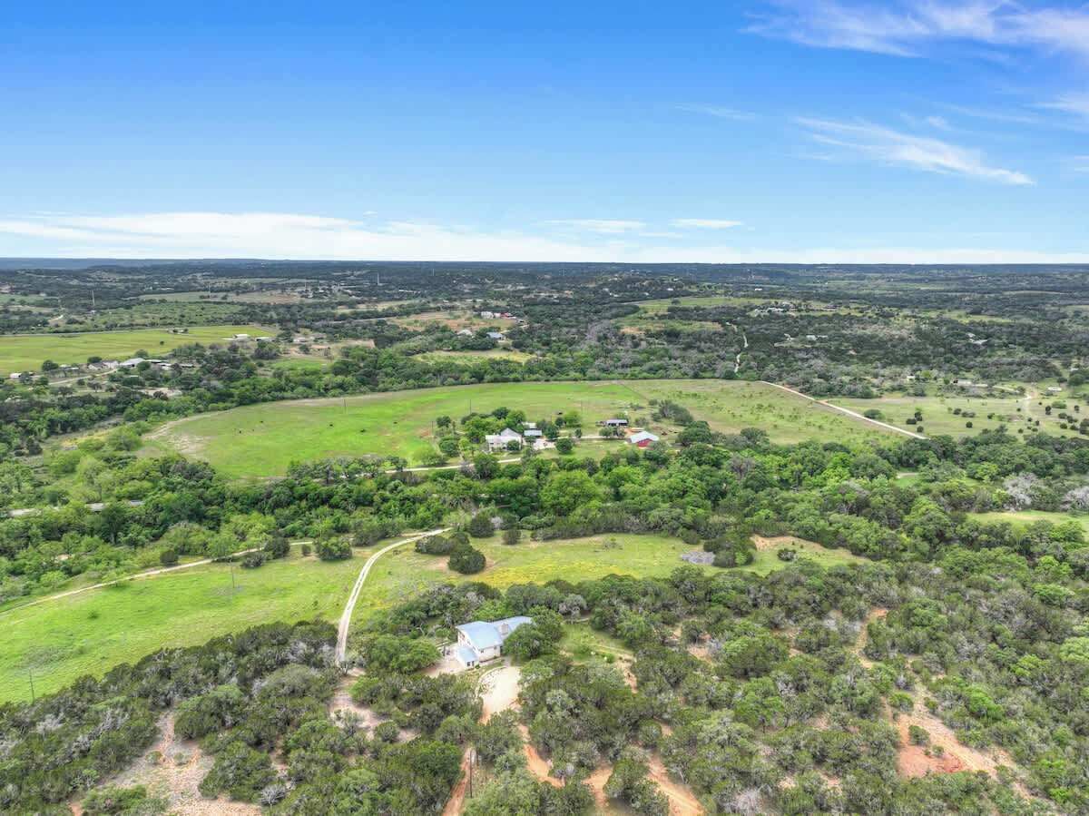 Vista aérea de un paisaje rural con edificios dispersos, campos verdes y un camino de tierra sinuoso.
