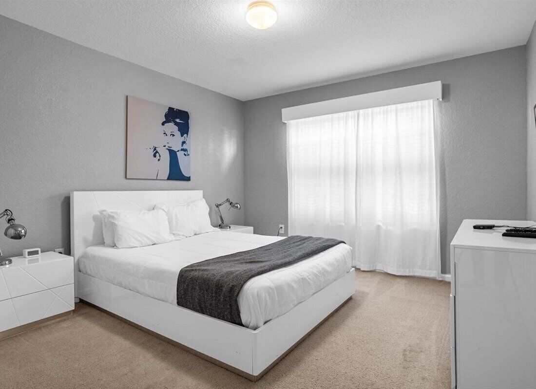 Un dormitorio moderno con cama blanca, una mesita de noche con lámpara, un escritorio con computadora y un amplio ventanal con cortinas translúcidas.