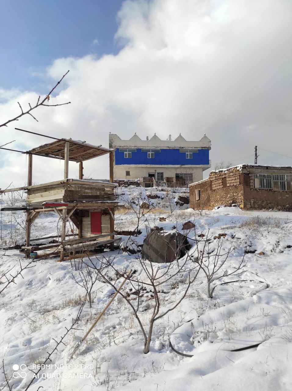 A snow-covered outdoor area with a blue building, a wooden structure, and a stone wall.