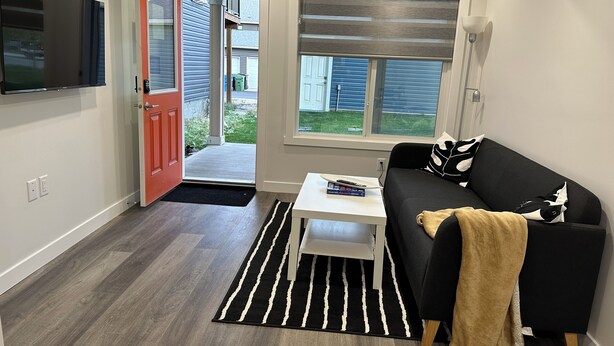 A compact living space with a sofa, a coffee table, a TV mounted on the wall, a sliding door, and a window with blinds.