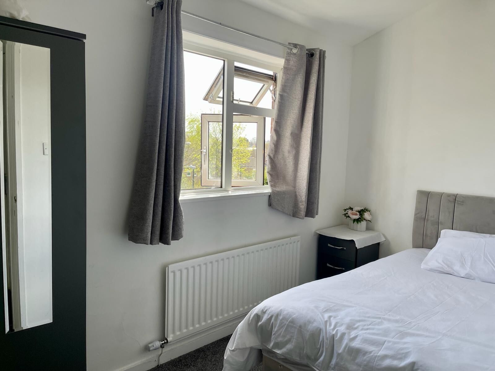 A bedroom with a bed, a radiator, a window with curtains, a bedside table with a vase, and a wardrobe.