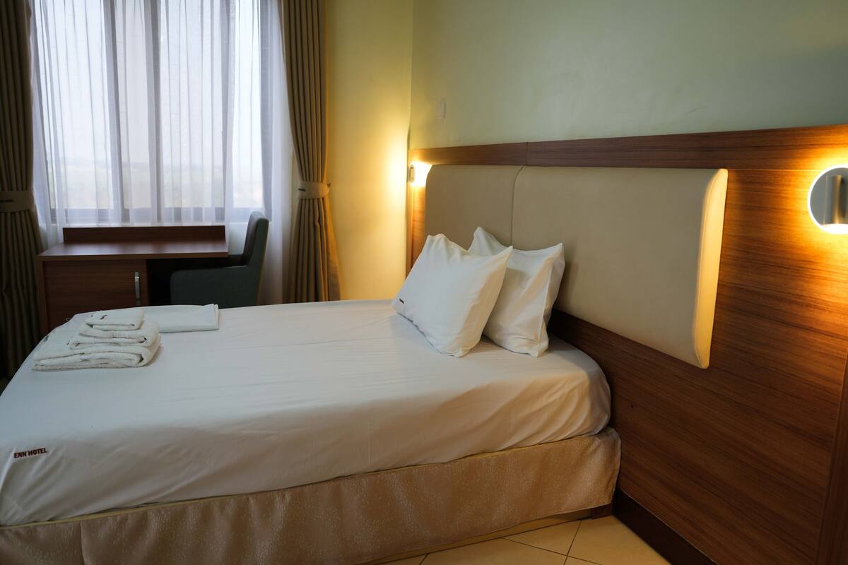 A neatly made bed with white linens and pillows, a wooden headboard with a light fixture, and a window with sheer curtains.
