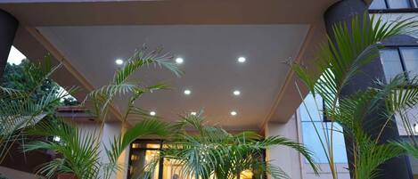 Entrada de un edificio con un patio techado, plantas en macetas y una puerta de vidrio que refleja a una persona en el interior.