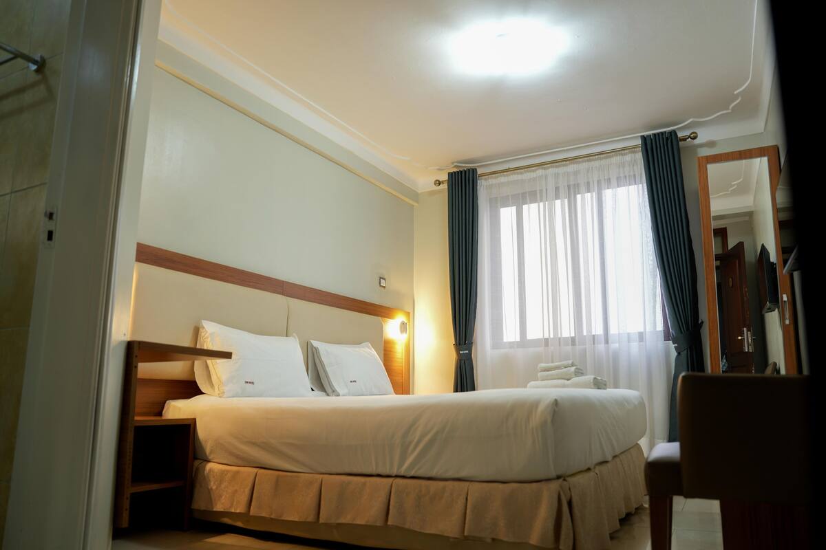 A hotel room with a bed, a nightstand, a chair, a mirror, and a window with curtains.