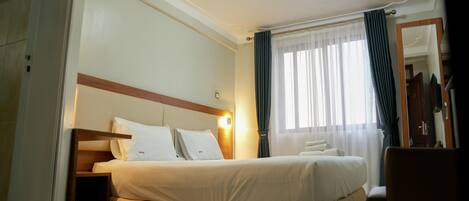 A hotel room with a bed, a nightstand, a chair, a mirror, and a window with curtains.
