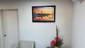 Una habitación con un sillón de color claro, otro sillón más oscuro, una mesita con una planta decorativa, un cuadro con una vista urbana enmarcada y un equipo de aire acondicionado en la pared.