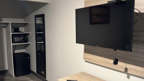Una habitación moderna con una televisión de pantalla plana montada en una pared de madera, un escritorio con una silla, un microondas, un refrigerador pequeño y un basurero.