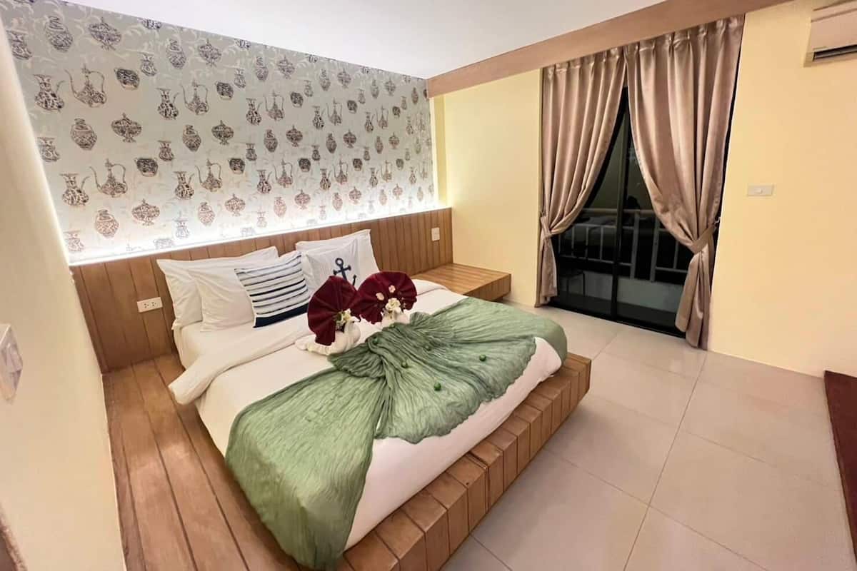 A modern bedroom with a wooden headboard, a large bed with a green throw, white bedding, and a patterned wallpaper.