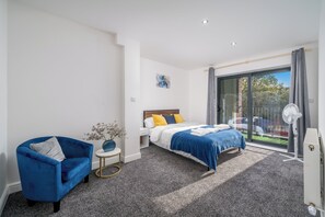 Een slaapkamer met een bed, een blauwe fauteuil, een klein bijzettafeltje, een ventilator en uitzicht op buiten.
