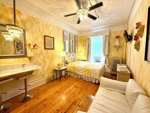 A hotel room with a wooden floor, a bed with a yellow and white bedspread, a ceiling fan, a large mirror, a vanity with a marble countertop, and a window with curtains. - Chic 2-Room Studio Retreat, Quiet Comfort in New York (New York)