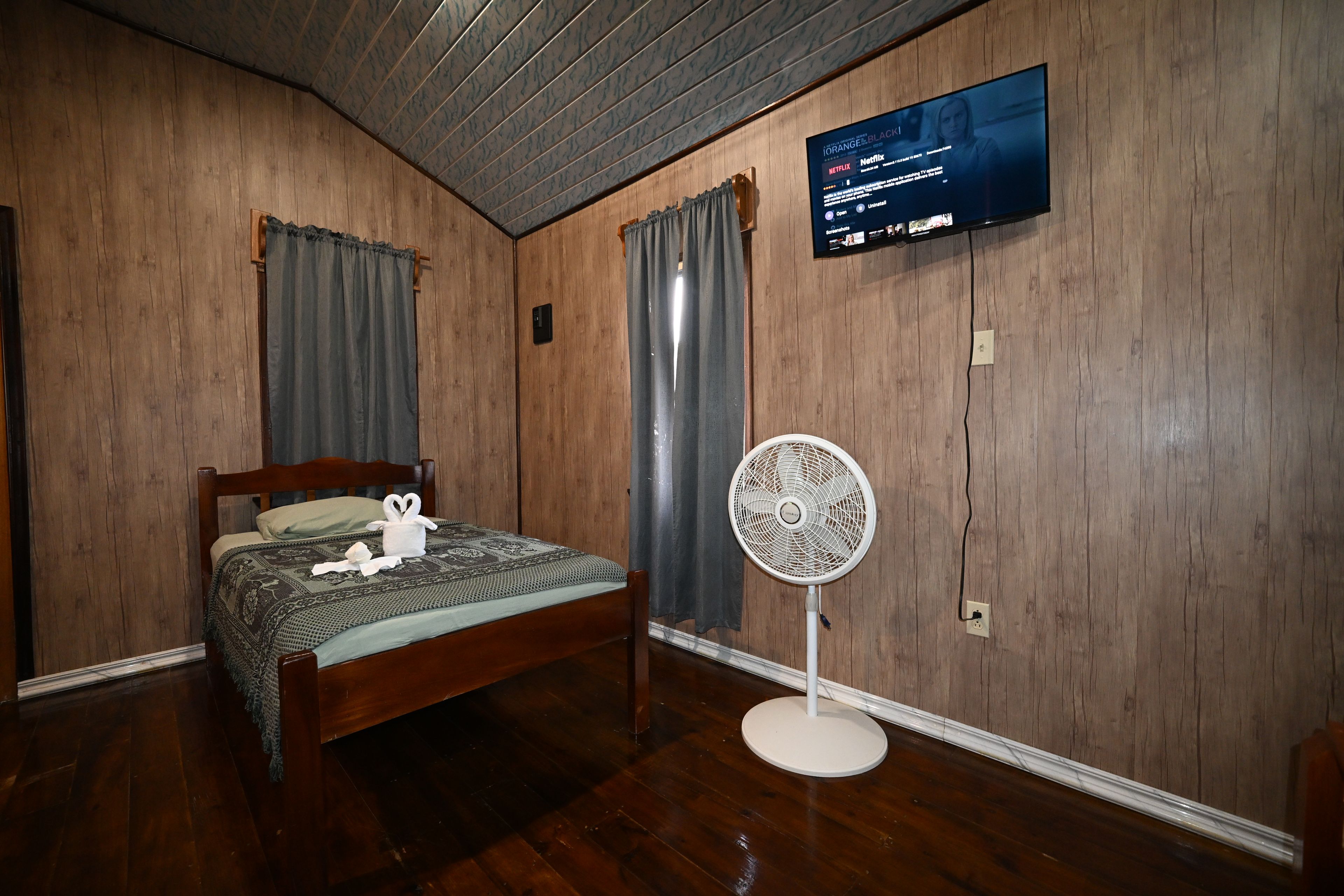 Une pièce avec un plancher en bois, un lit recouvert d’une couvre-lit à motifs, un téléviseur fixé au mur et un ventilateur sur pied.