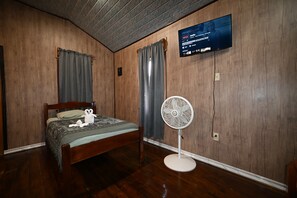 Una habitación con piso de madera, una cama con colcha estampada, un televisor montado en la pared y un ventilador de pie.