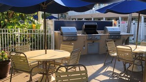 An outdoor patio with a grill, tables, and chairs under blue umbrellas.