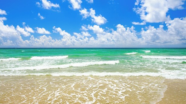A beach with clear turquoise water and white foam waves, under a bright blue sky with scattered clouds.