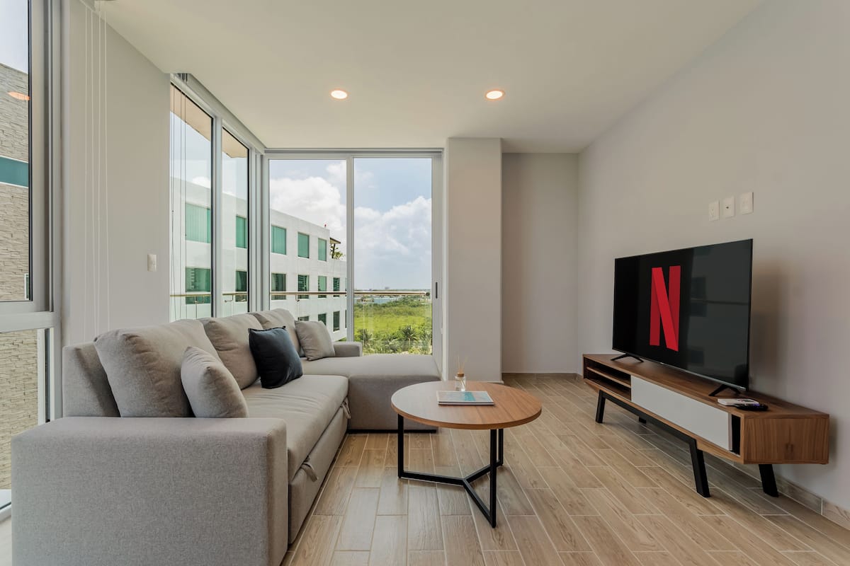 Ein modernes Wohnzimmer mit einer Couch, einem runden Couchtisch, einem Fernseher und großen Fenstern mit Blick auf Gebäude und Grünflächen.