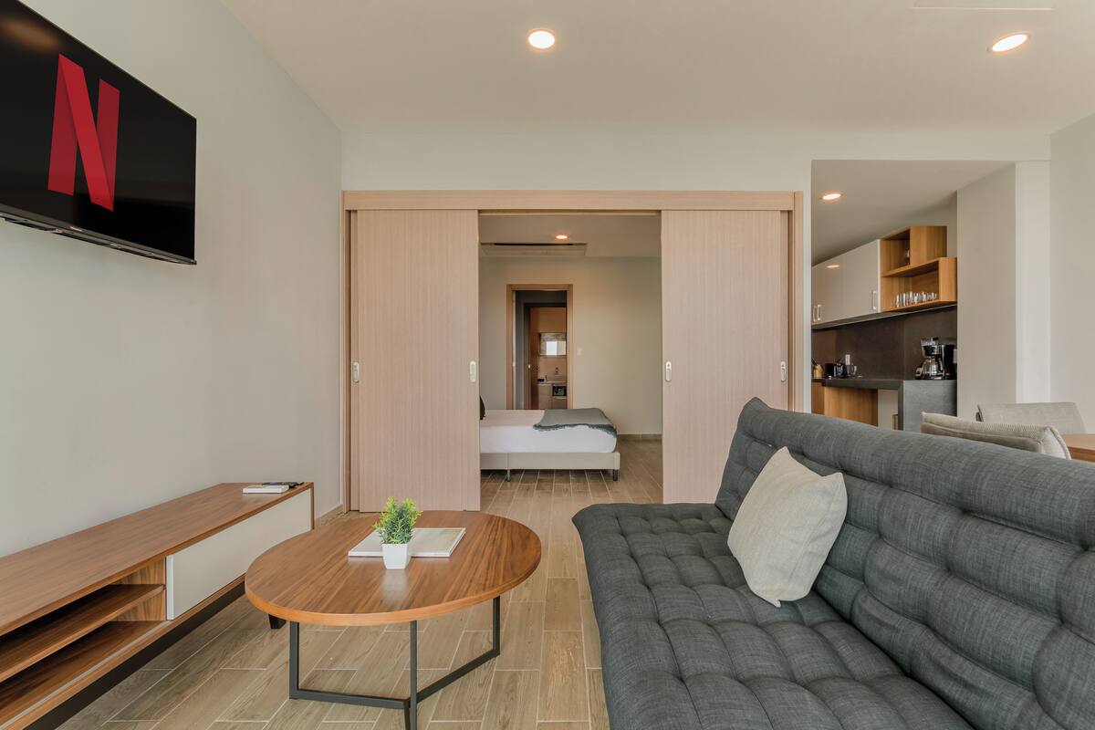 Ein modernes Hotelzimmer mit grauem Sofa, einem Holztisch, einem Fernseher mit Netflix-Logo, einem Bett und einer Küchenzeile.