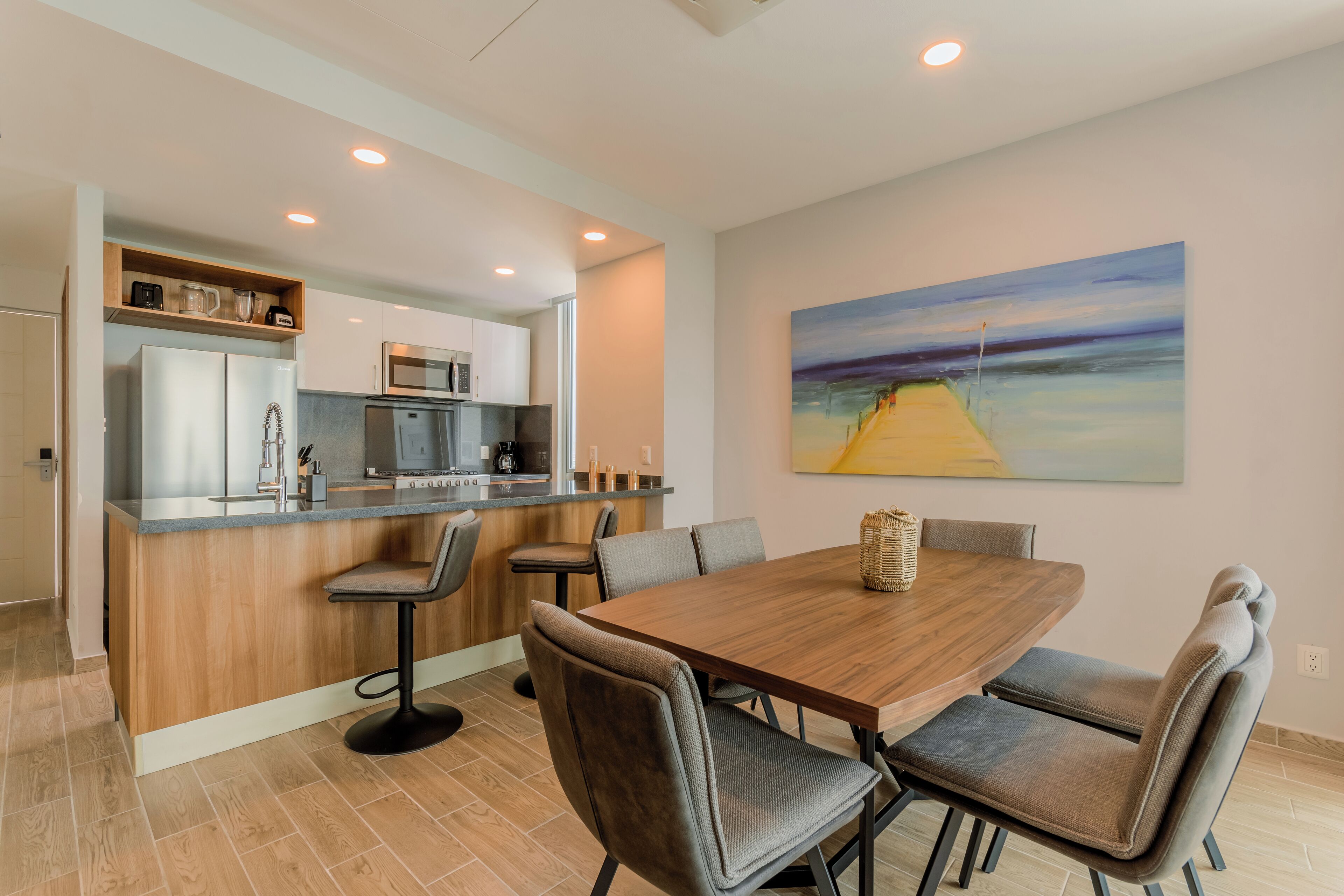 Una cocina moderna con una isla central, una zona de comedor con una mesa y sillas de madera, y un cuadro grande en la pared.