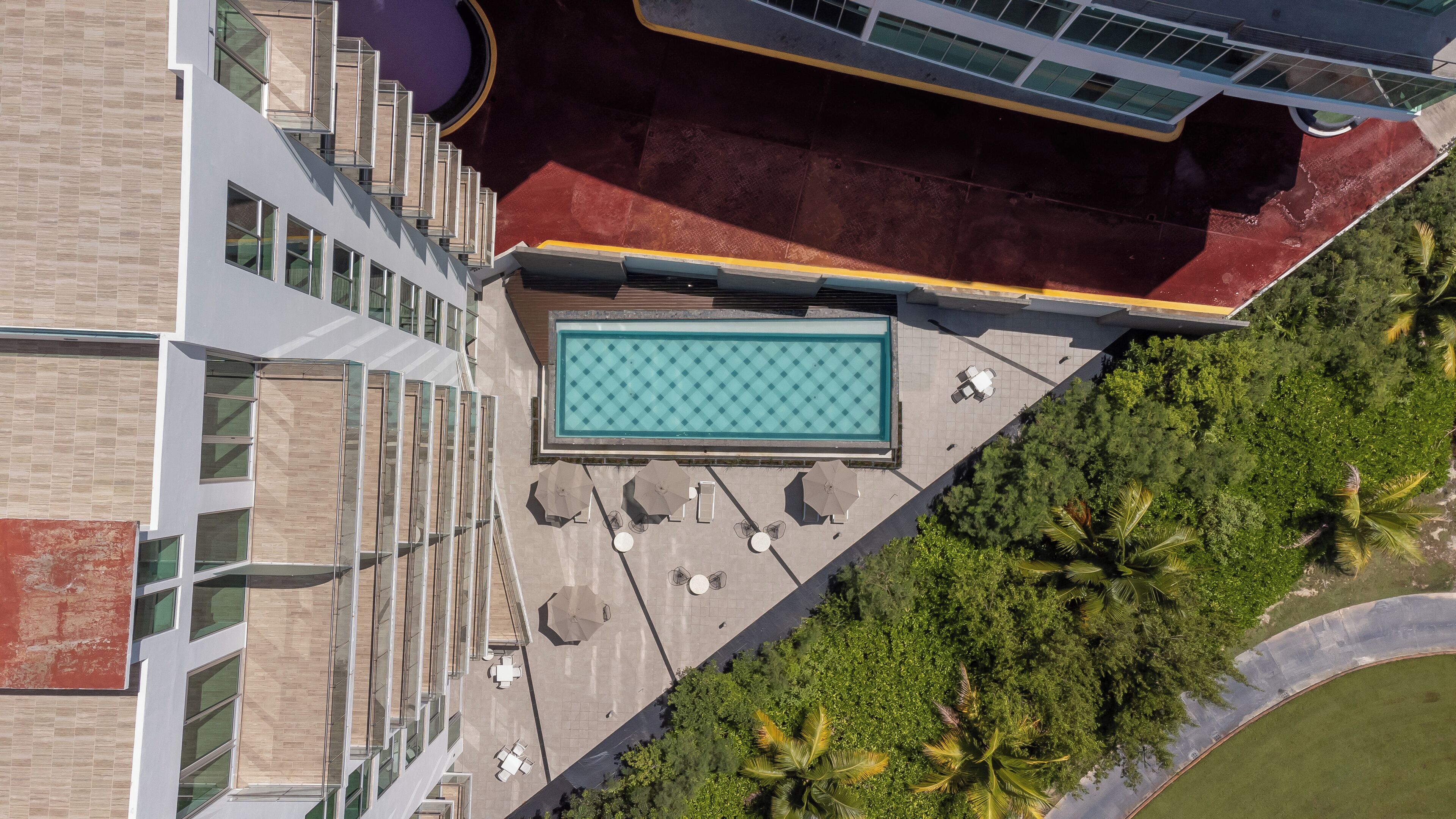 An aerial view of a modern building with a rooftop pool, surrounded by lush greenery and palm trees.