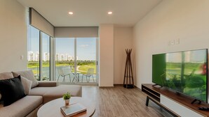 Een moderne woonkamer met een grote televisie, een bank, een salontafel met een boek en een plant, en een balkon met uitzicht op een golfbaan.