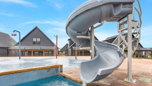 A water slide structure over a pool with a clear sky and residential buildings in the background. - Solid Rock Lodge >Lake View >Hot Tub >Indoor Pool > Near Big Cedar&Thunder Ridge (Hollister)