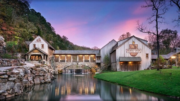 A rustic lodge with a stone bridge over a reflective water body, surrounded by lush greenery and a mountainous backdrop. - Solid Rock Lodge >Lake View >Hot Tub >Indoor Pool > Near Big Cedar&Thunder Ridge (Hollister)