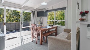 Ein geräumiges Zimmer mit einem Holzesstisch, einer Couch und einem Fernseher. Große Fenster bieten einen Blick ins Freie.