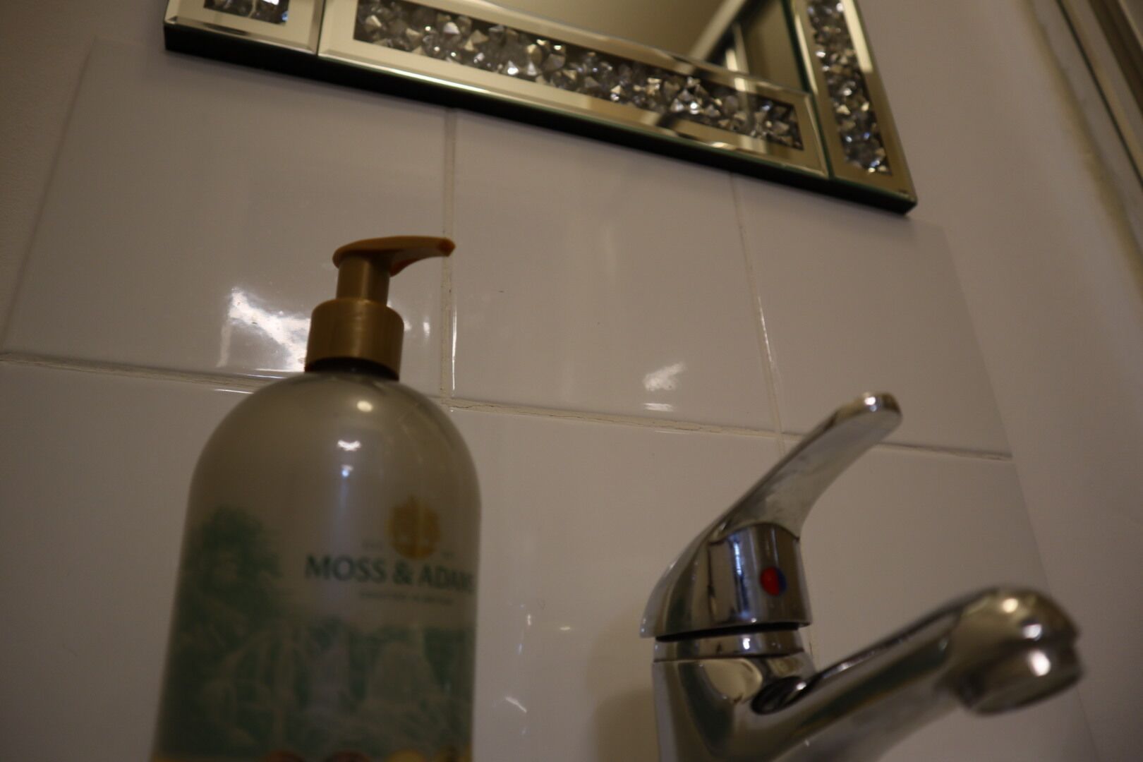 A bathroom with a white tiled wall, a chrome faucet, and a bottle of "Moss & Adam" shampoo.