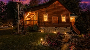 A wooden cabin with a stone foundation, a porch with string lights, and a stone staircase leading to the entrance.