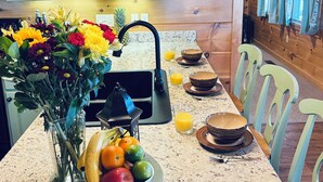 A kitchen with light green cabinets, a granite countertop, a fruit bowl, a vase of flowers, and a dining table set for breakfast.