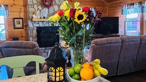 A cozy cabin interior with a stone fireplace, a ceiling fan, a dining table with a lantern, and a bowl of fruit.