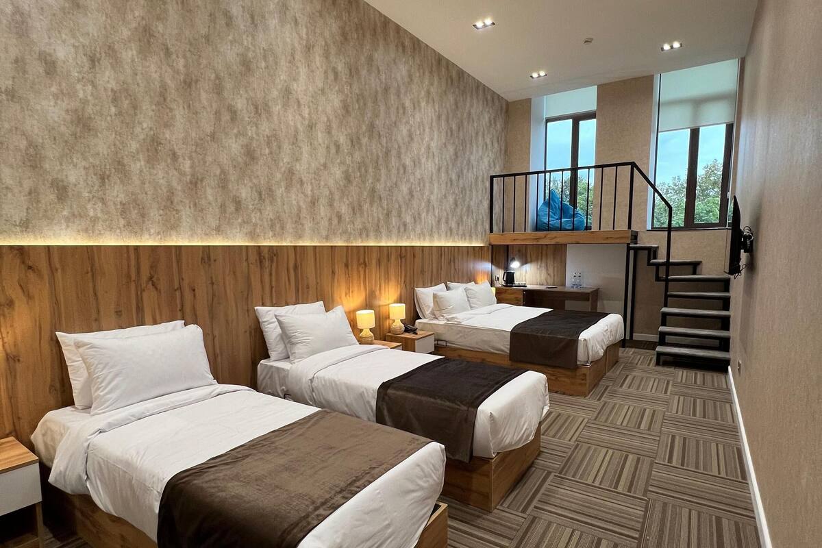 A hotel room with three beds, wooden headboards, a staircase, and large windows.