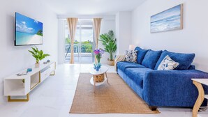 A modern living room with a blue sofa, a flat-screen TV, and a large window with curtains. - Modern 1BR Apt in Hollywood, FL – 5 Mins to Beach, W/ Pool, Gym, & Free Parking (Hollywood)