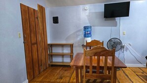 Ein Zimmer mit einem Holztisch, Stühlen, einem Wasserspender, einem Ventilator und einem an der Wand befestigten Fernseher.