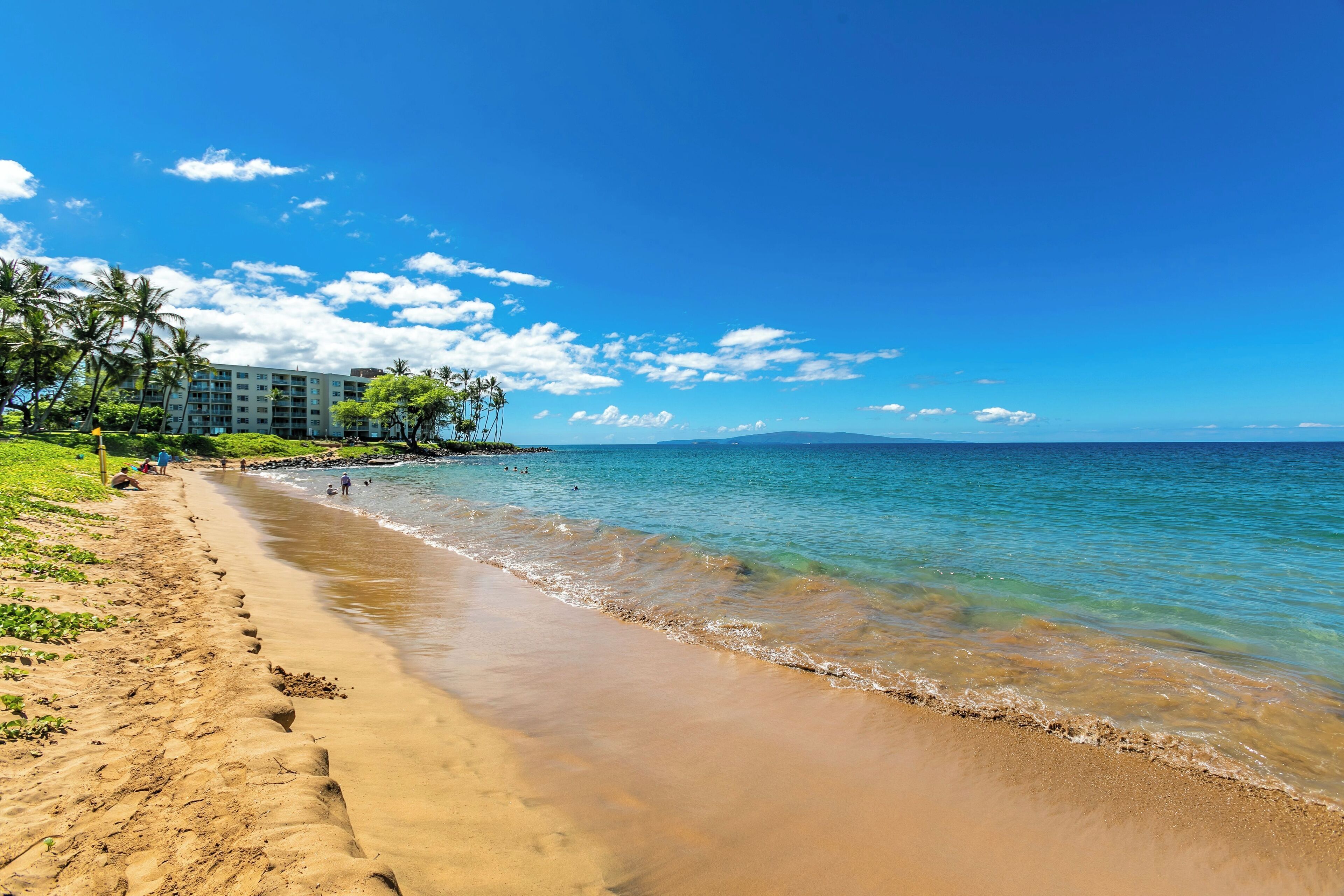 A beach with clear blue water, a sandy shore, and a building with a pool area.