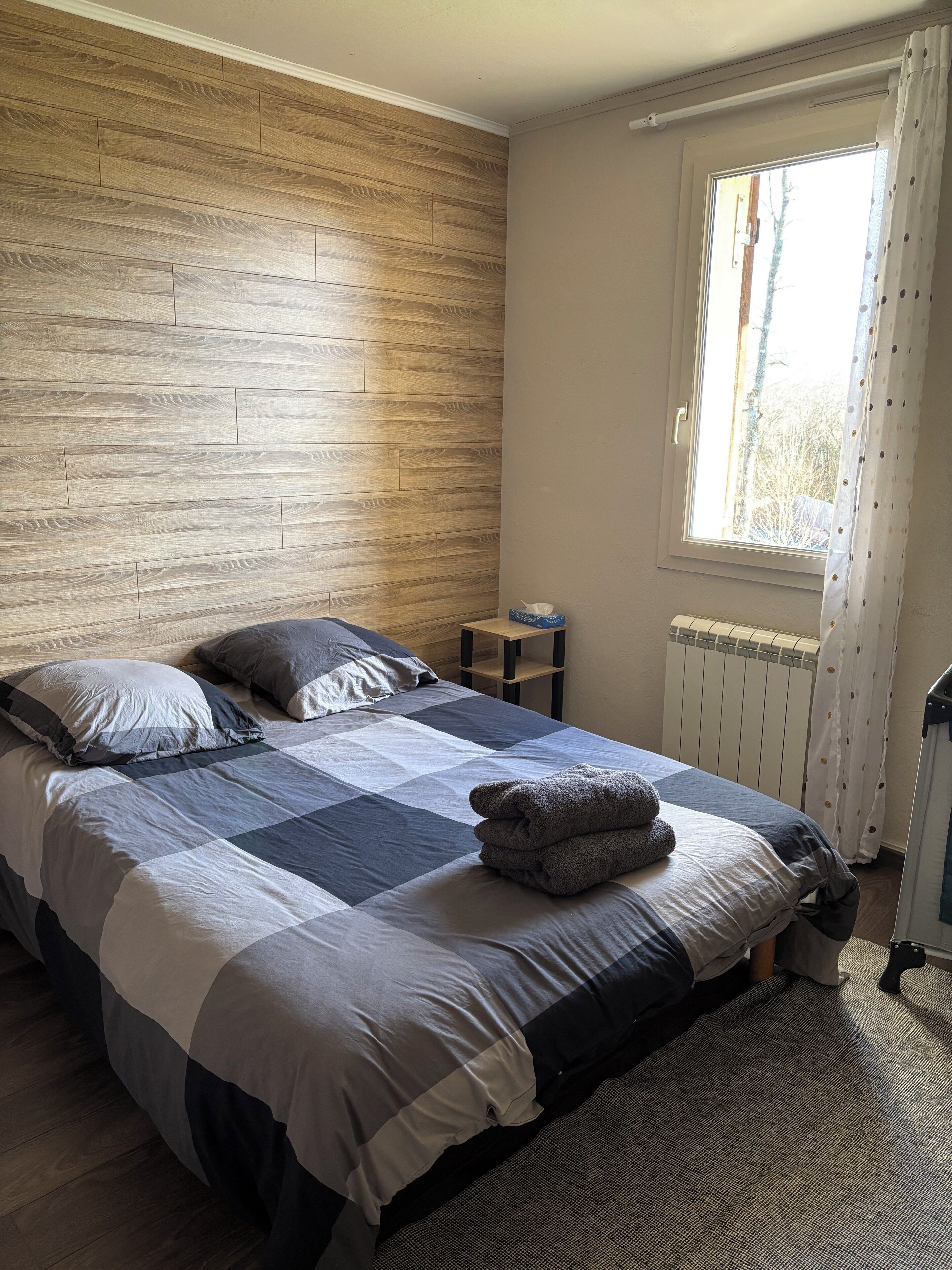Ein Schlafzimmer mit Holzkopfende, einem Bett mit Patchworkdecke, einem Nachttisch, einem Heizkörper und einem Fenster mit Vorhängen.