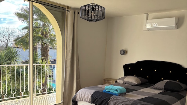 Une chambre à coucher avec un lit, un ventilateur de plafond, une fenêtre donnant sur des palmiers et une porte-fenêtre coulissante.