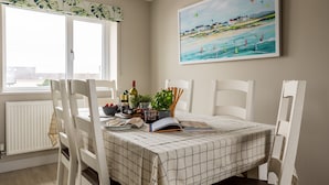 A dining area with a table set for four, white chairs, a checkered tablecloth, a potted plant, a wine bottle, and a book. - Awelfa: Modern 3-Bed Home in Rhosneigr | Managed by Anglesey Holiday Lettings (Rhosneigr)