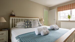A neatly made bed with a blue patterned bedspread, two pillows, and rolled towels. There is a nightstand with a lamp and a book. A window with a striped valance and a potted plant is visible. - Awelfa: Modern 3-Bed Home in Rhosneigr | Managed by Anglesey Holiday Lettings (Rhosneigr)