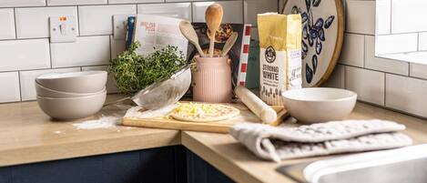 A kitchen counter with a pizza, bowls, a rolling pin, and a recipe book.