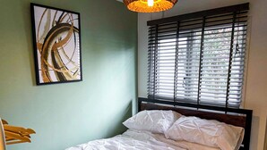 Une chambre à coucher avec un lit, une fenêtre avec des stores, un luminaire suspendu et un tableau abstrait encadré accroché au mur.