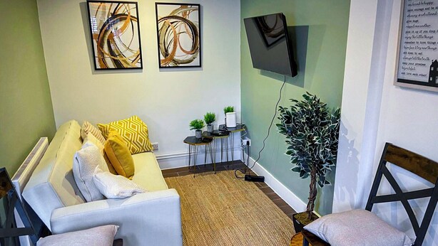 Un salon comprenant un canapé, deux fauteuils, une petite table avec une plante et un téléviseur fixé au mur.